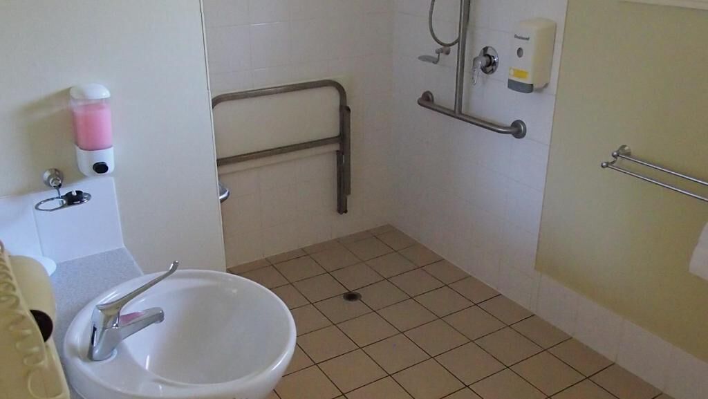 Baño con lavamanos, dispensador de gel antibacterial, barra de apoyo y zona de ducha con dispensador de jabón.