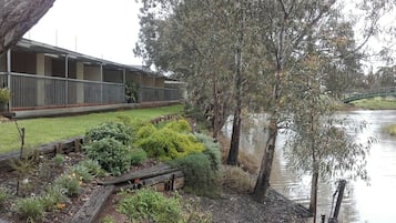 A riverside view of a building with a covered outdoor area, a wooden deck, and a bridge in the background.