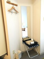Habitación de hotel con un espejo grande, una percha de madera, un carrito de equipaje negro y un cubo de basura blanco.