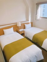 Habitación de hotel con dos camas, una mesita de noche, una lámpara y un ventanal con cortinas.