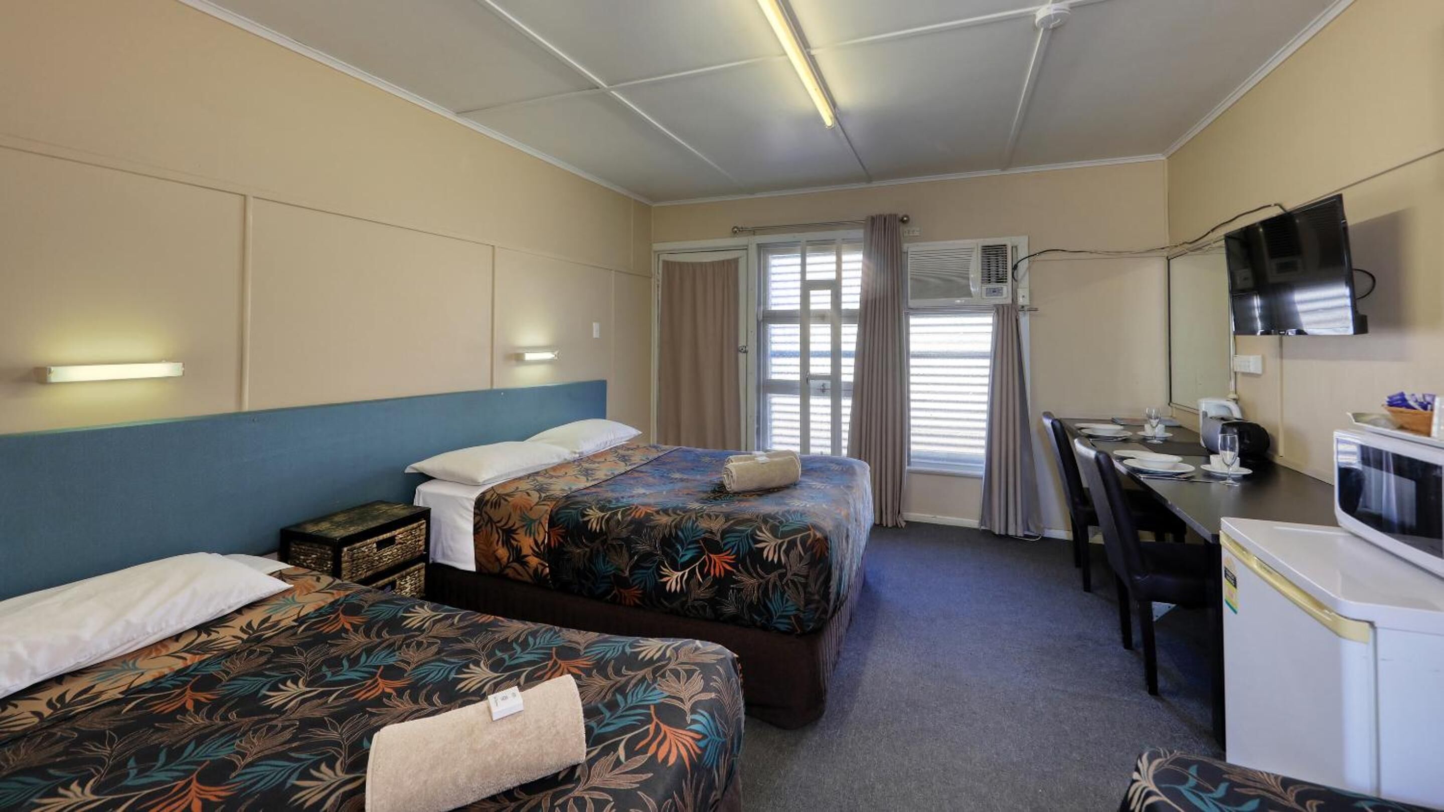 A hotel room with two beds, a desk, a chair, a television, and an air conditioning unit.