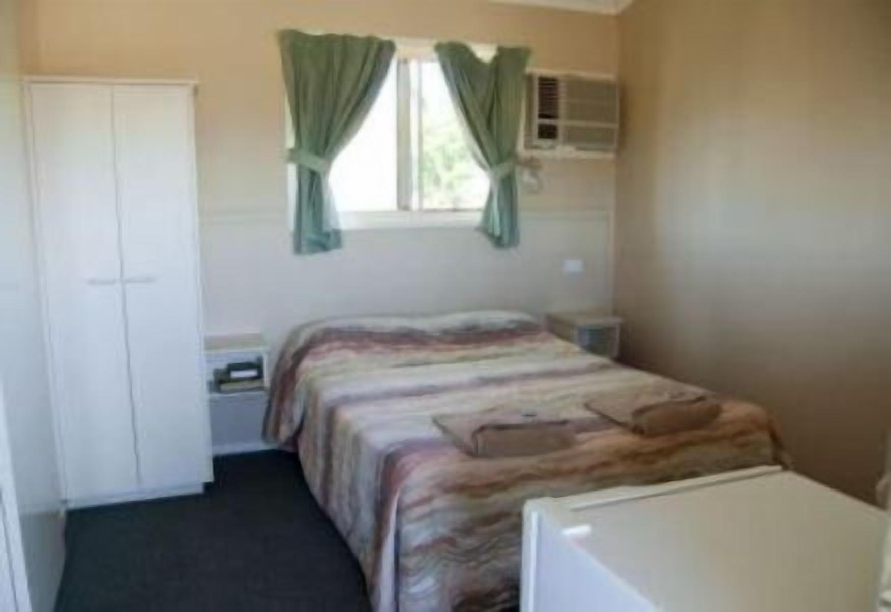 A small, single-bed room with a bed, a wardrobe, a window with curtains, a bedside table, and a small heater.