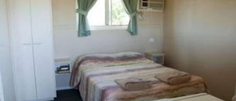 A small, single-bed room with a bed, a wardrobe, a window with curtains, a bedside table, and a small heater.