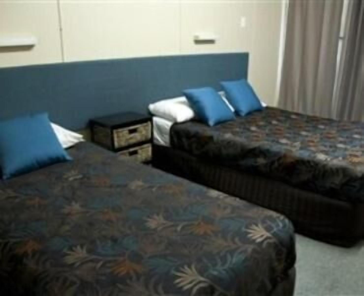 Habitación de hotel con dos camas, cada una con una colcha estampada y almohadas azules. Entre las camas, hay una mesita de noche con una lámpara.