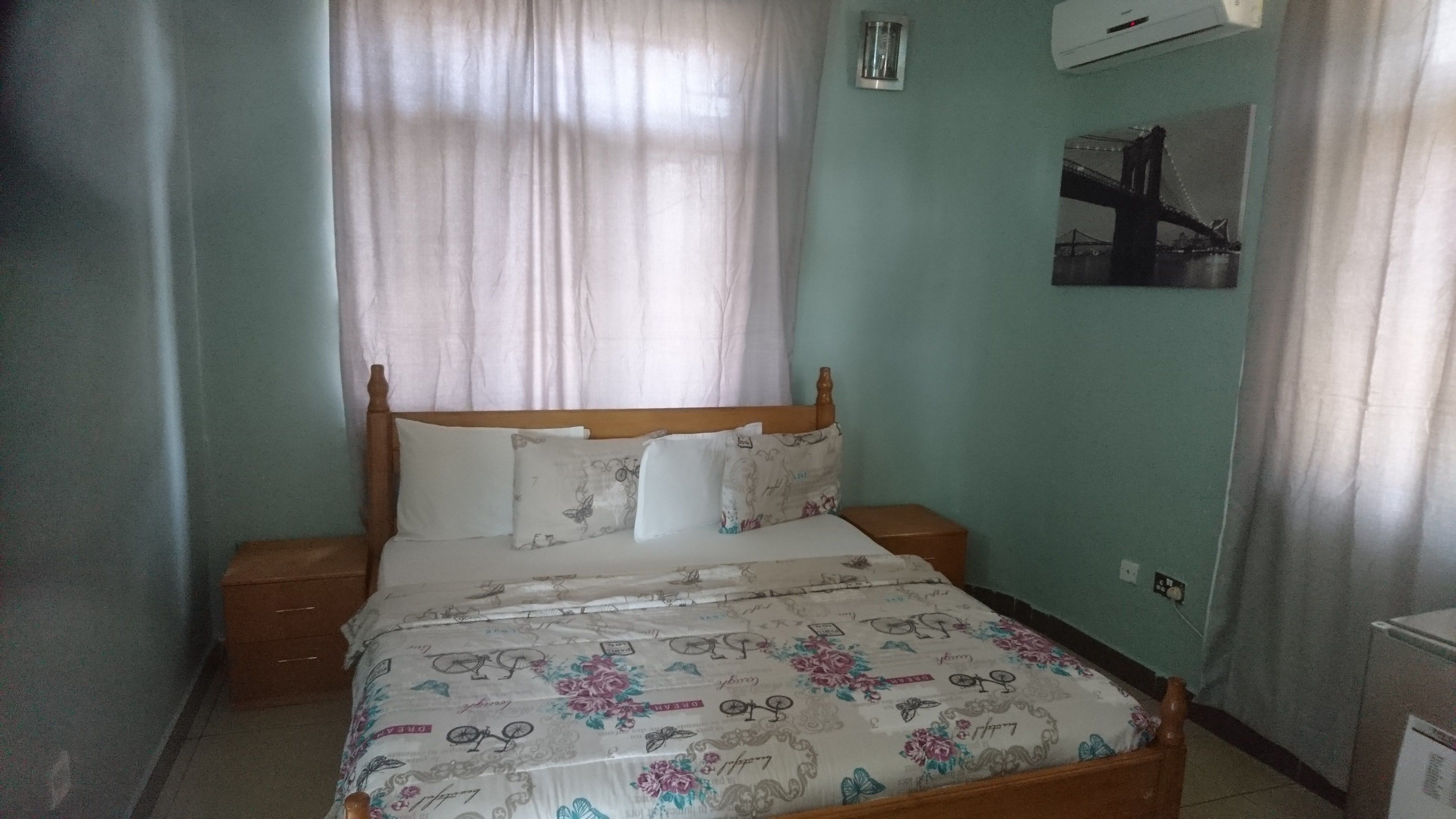 Quarto com cama de madeira, roupa de cama branca, mesa de cabeceira, ar condicionado fixado na parede e um quadro emoldurado.