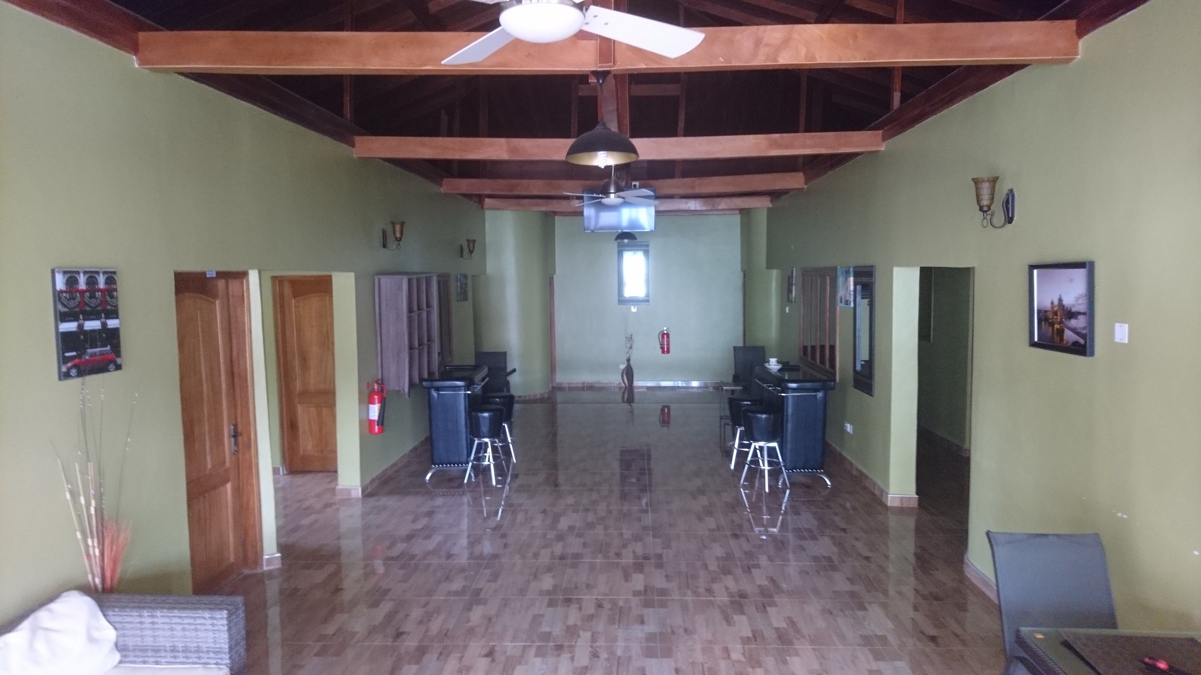 A spacious hallway with a high ceiling, wooden beams, and a ceiling fan. There are two bar areas with stools and a fire extinguisher on the wall.