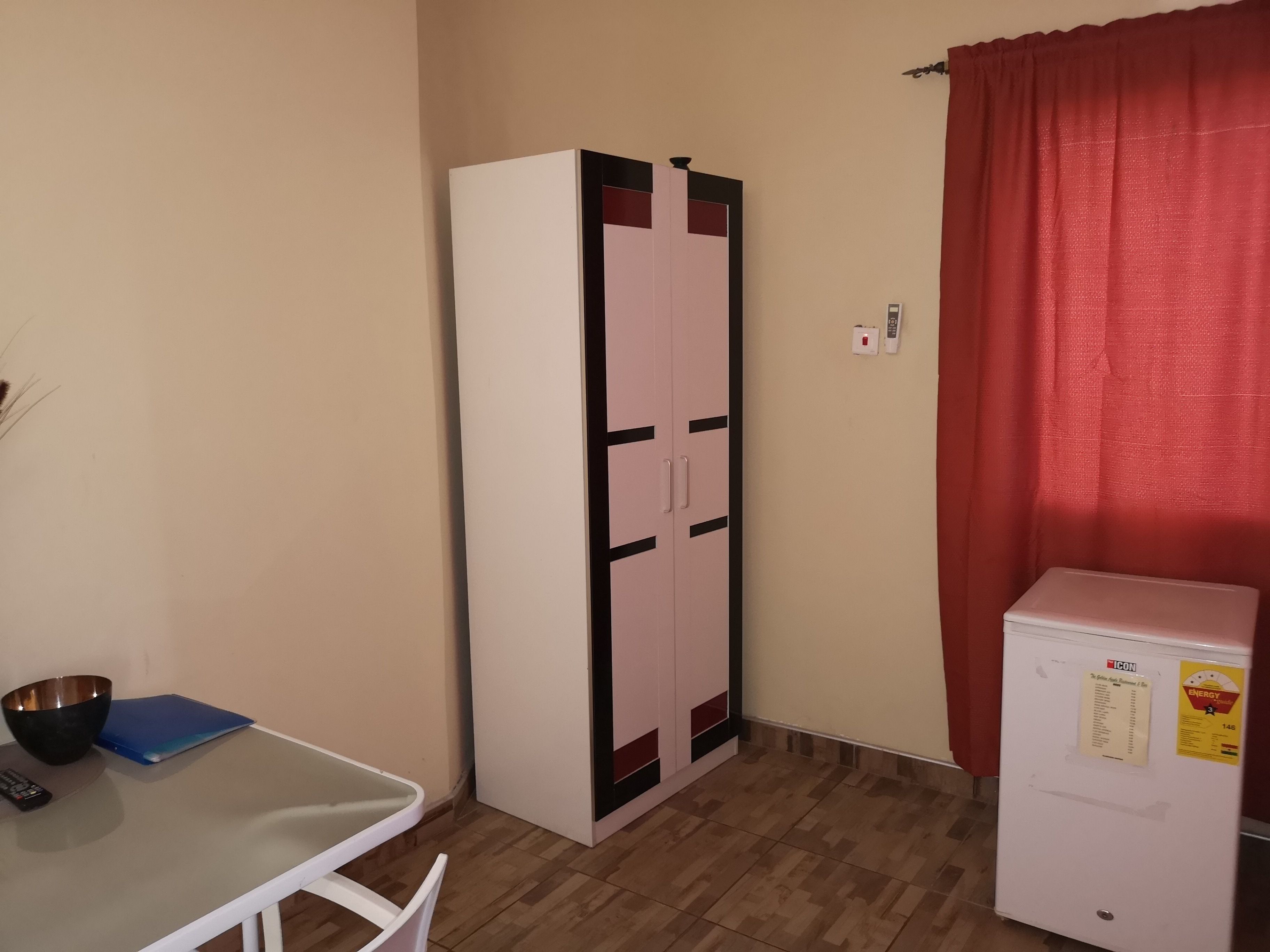Una habitación pequeña con un armario blanco, un refrigerador blanco compacto, una mesa con una carpeta azul y una cortina roja.
