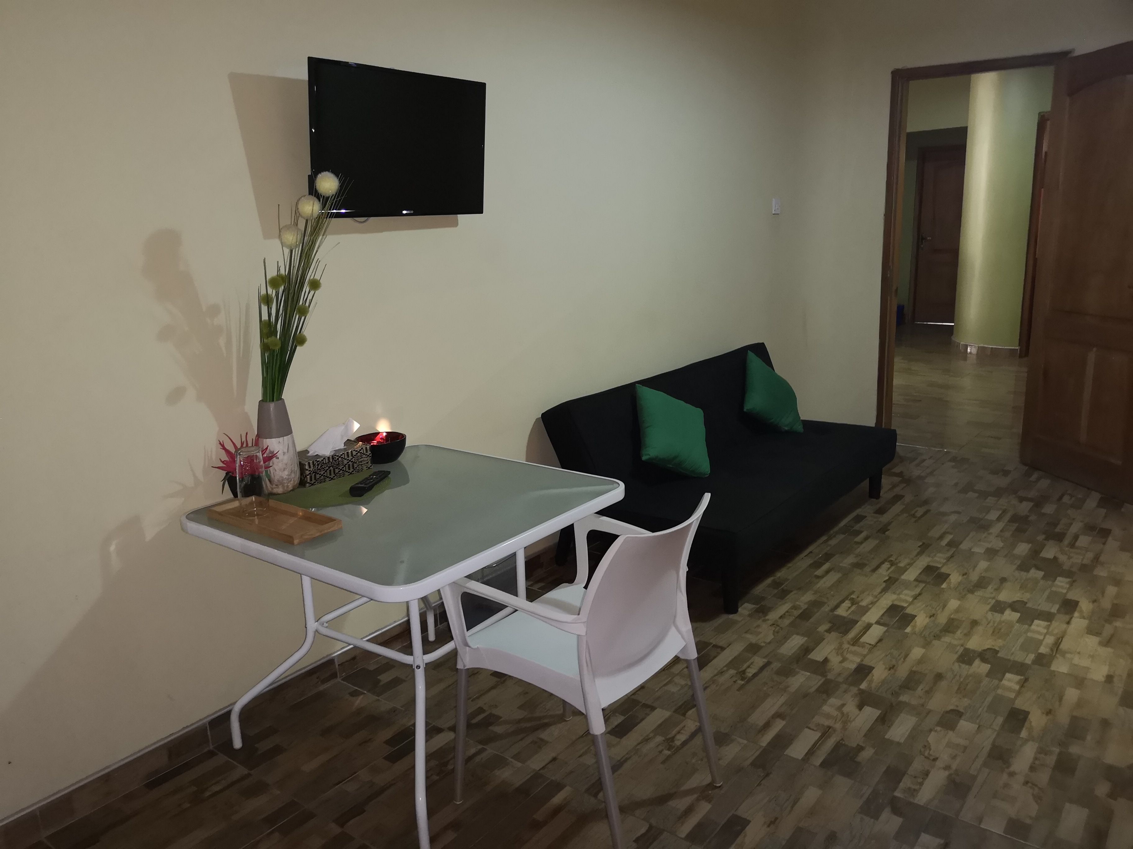 A small living area with a glass-top table, a white chair, a black sofa with green cushions, a flat-screen TV mounted on the wall, and a wooden floor.