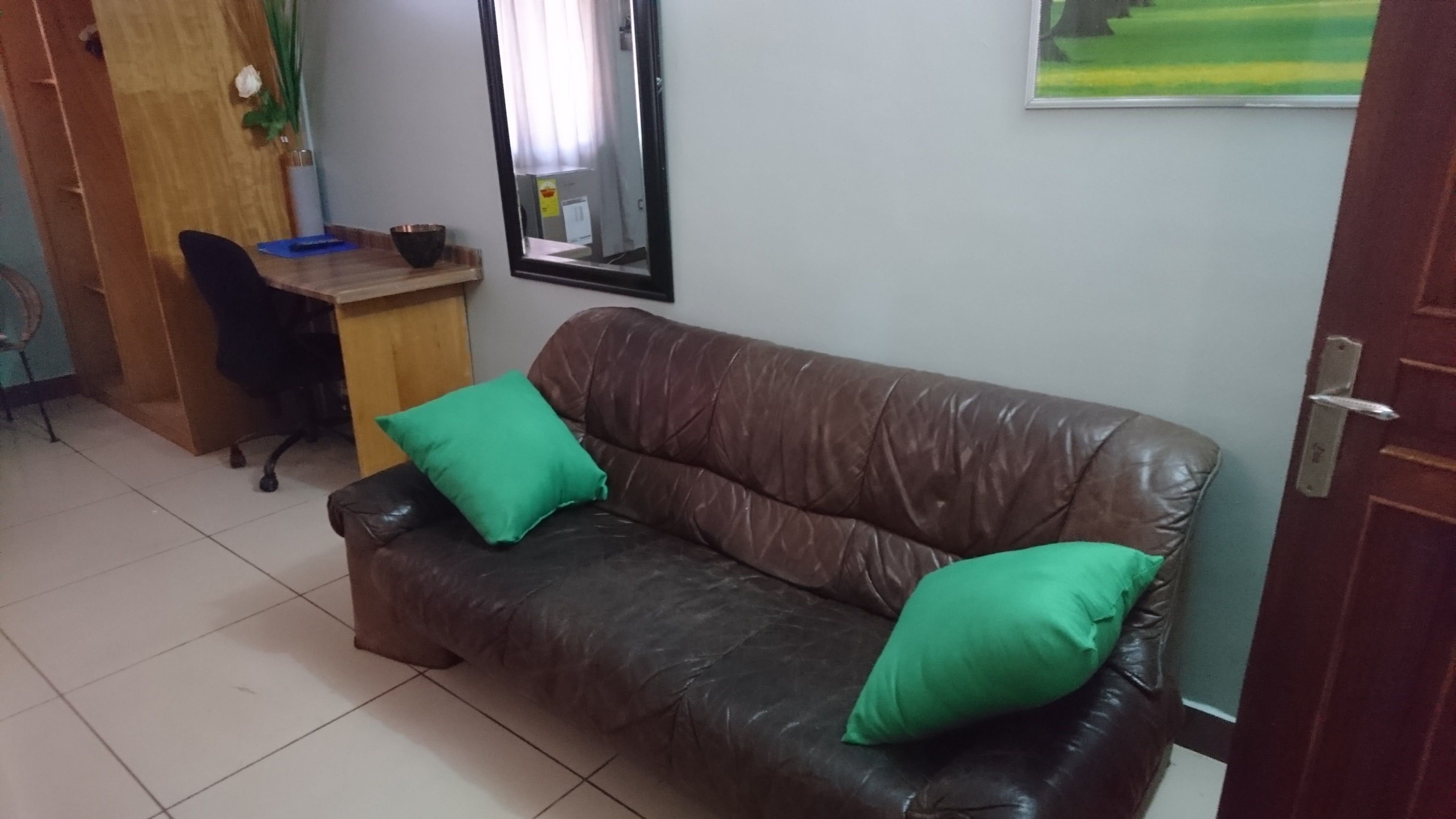 A room with a brown leather sofa, two green cushions, a wooden desk, and a chair.