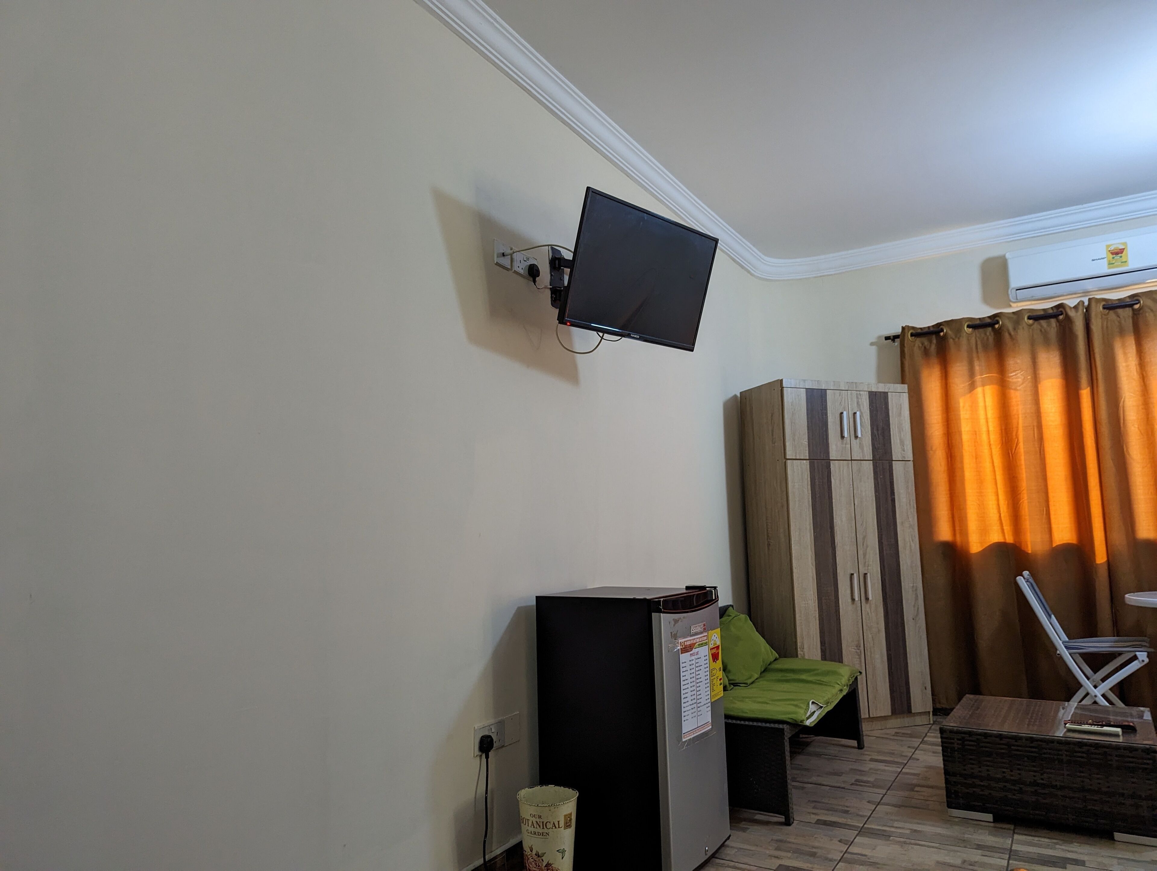 Habitació amb televisió de pantalla plana penjada a la paret, nevera petita, una banca amb coixins verds, armari de fusta i finestra amb cortines taronges.