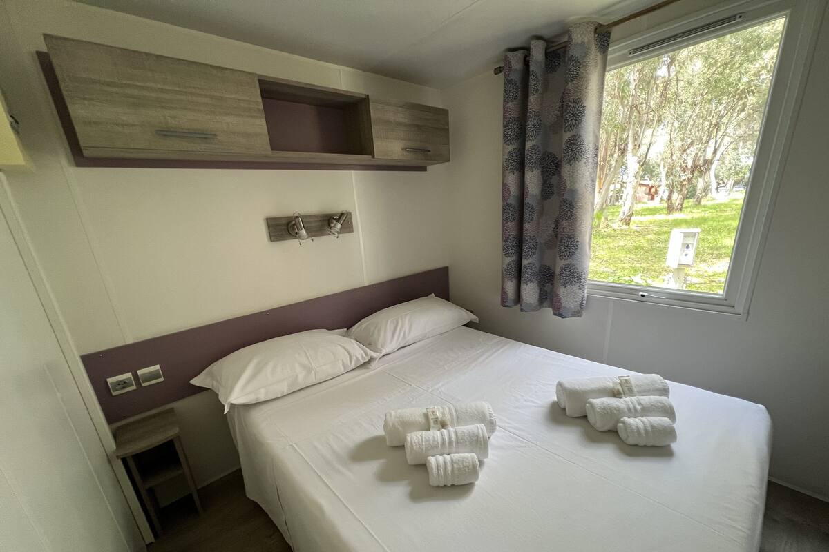 Een net opgemaakt bed met witte lakens en handdoeken, een nachtkastje met een lamp en een raam met gordijnen.