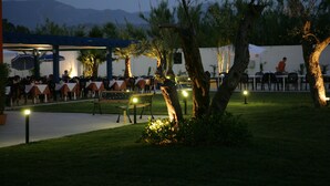 An outdoor dining area with tables and chairs, surrounded by trees and a building in the background. - Batuca Village & Camping (Terme Vigliatore)