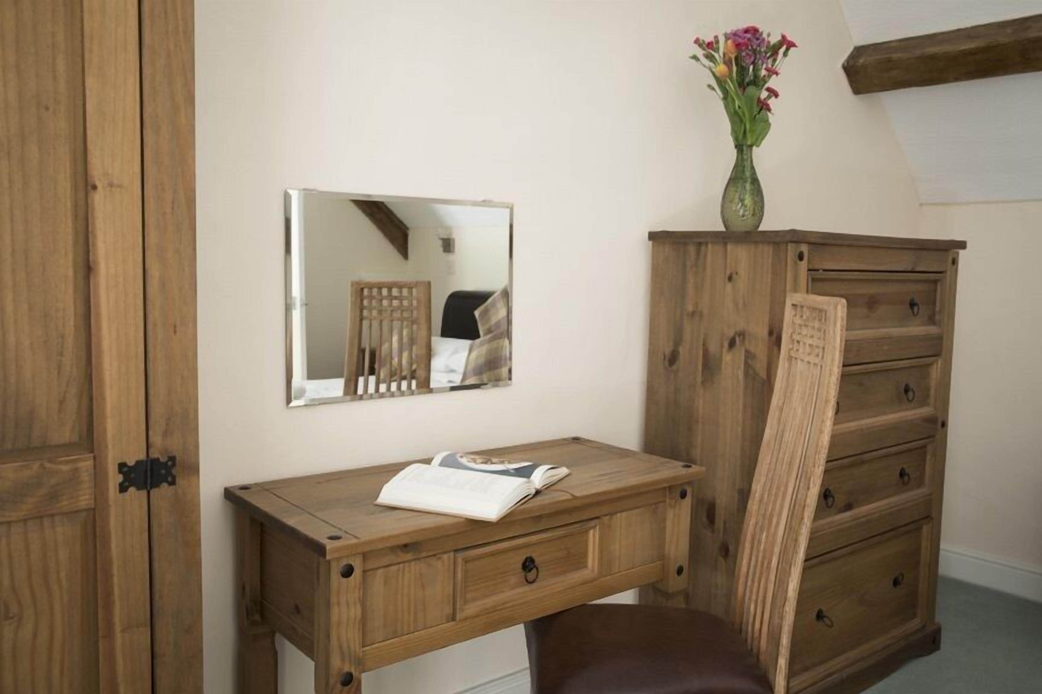 A wooden desk with a mirror, a vase with flowers, and a brown chair.