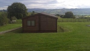 A small wooden cabin with a single window, surrounded by a grassy field and a wooden fence. - Long Mountain Centre (Shrewsbury)