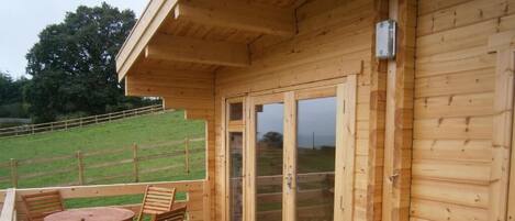 Um deck de madeira com mesa e cadeiras, uma porta de correr com vidro e vista para um campo gramado.