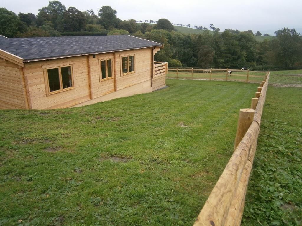 A wooden building with a sloped roof, a fenced area with grass, and a hilly landscape in the background.