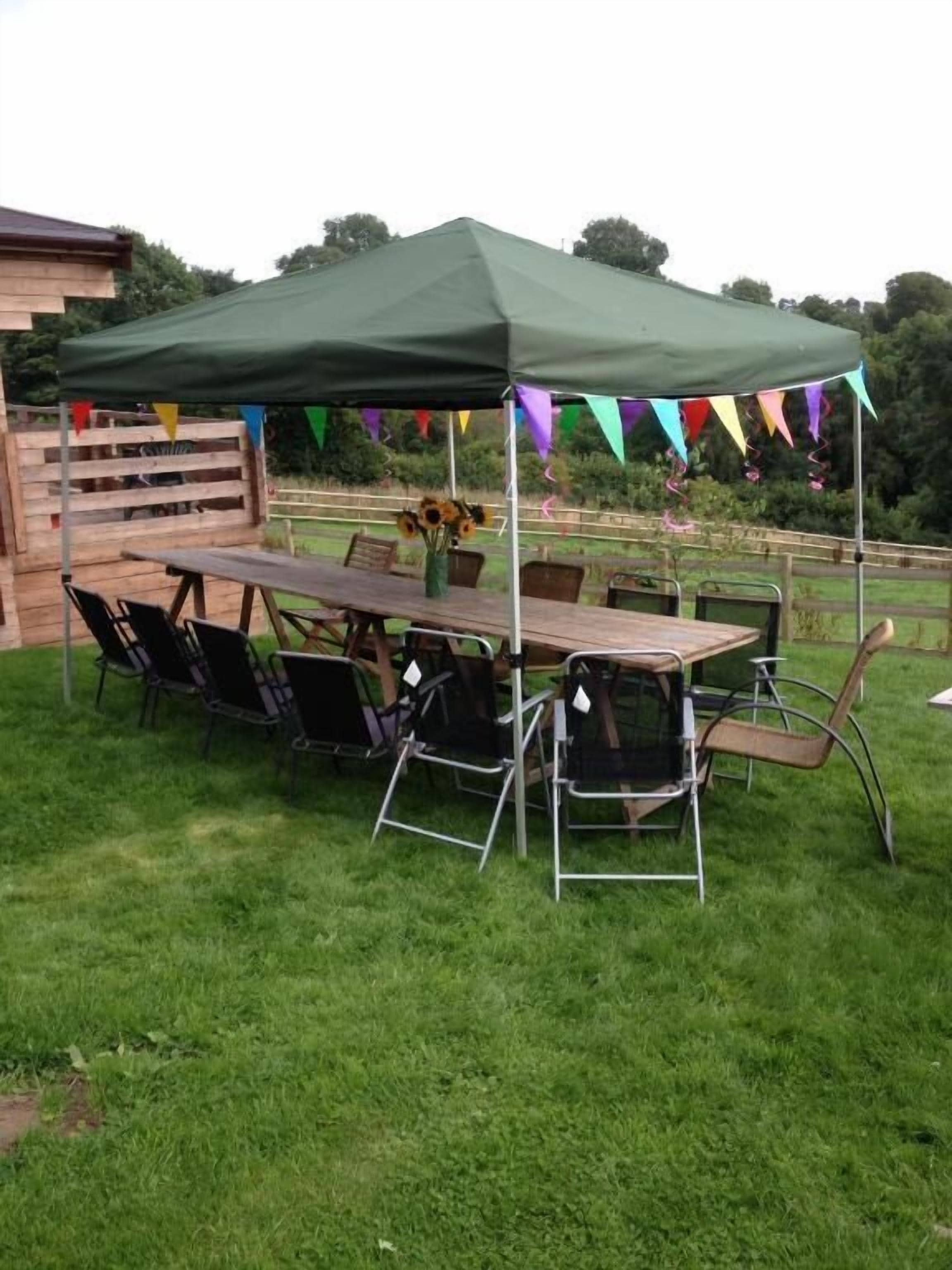 An outdoor dining area with a table and chairs under a green canopy, surrounded by a wooden fence and colorful flags.