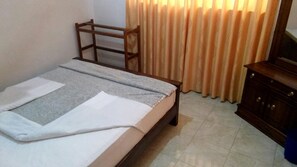 A single bed with a wooden frame, a grey and white bedspread, and a wooden headboard. There is a wooden dresser to the right and a curtain with a pattern in the background. - Amaya View Guest Inn (Haputhale)