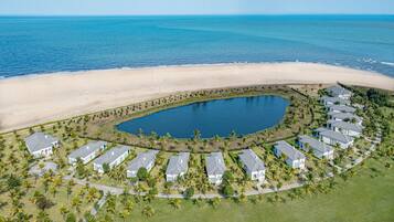 Vista aérea de um resort à beira-mar, com piscina circular, bangalôs na praia e palmeiras.