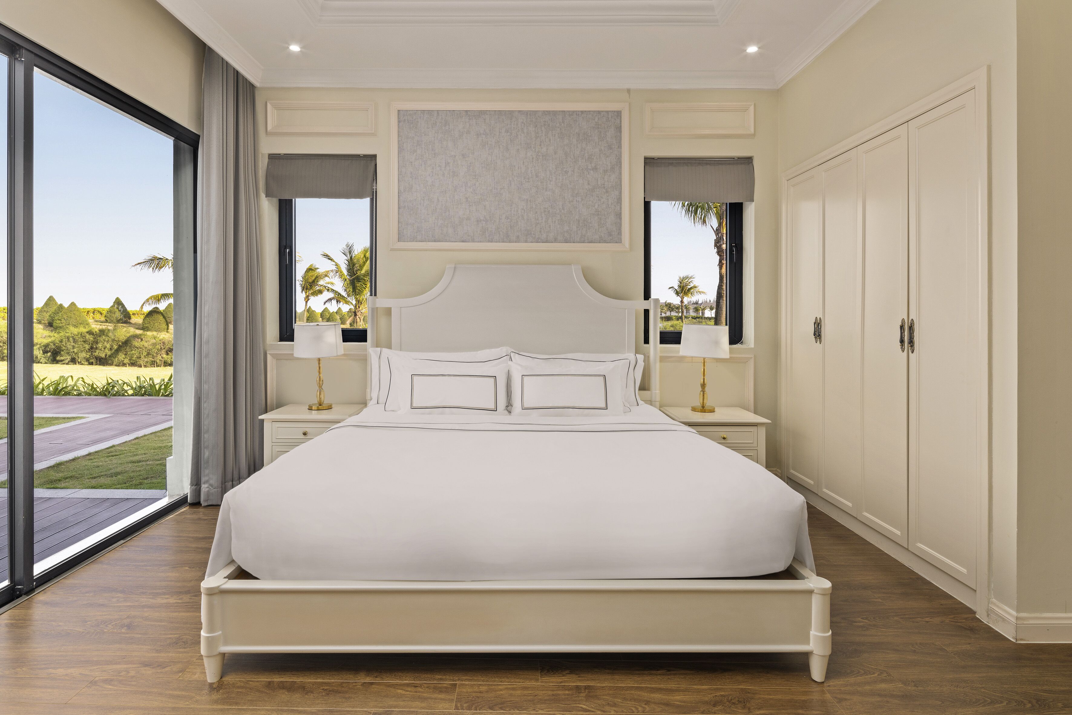 A large bed with a headboard, bedside tables, and a view of greenery outside.