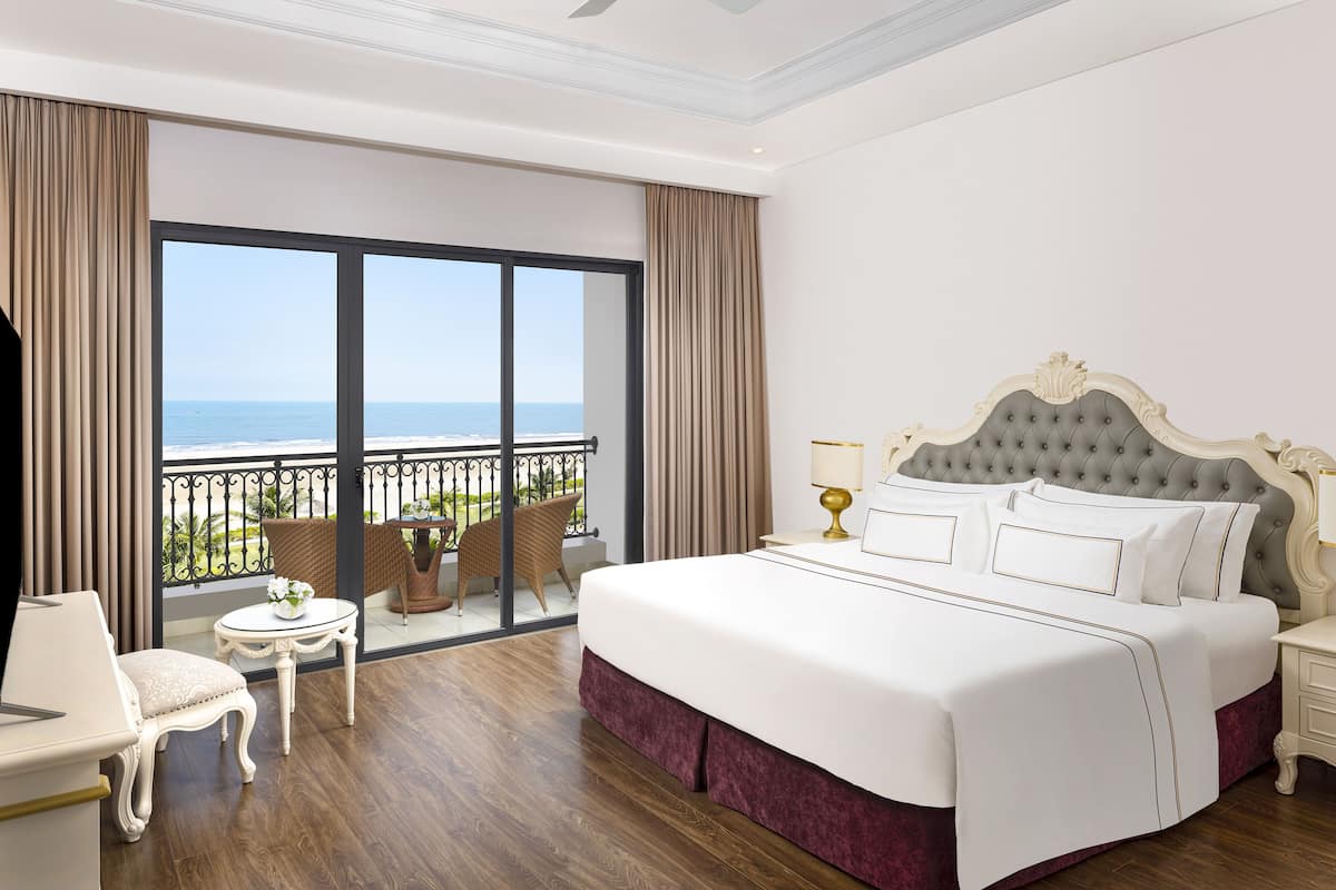 A hotel room with a large bed, a balcony with a view of the sea, a small table with a lamp, and a chair.