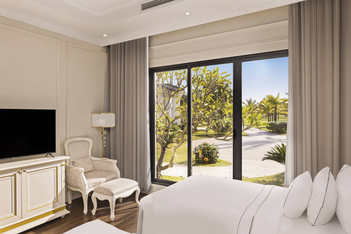 A hotel room with a large bed, a television, a seating area with a chair and ottoman, and a view of a tropical landscape through large windows.