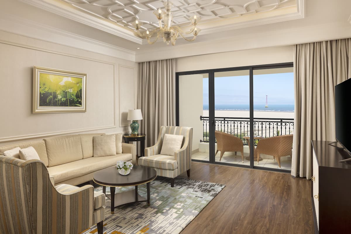 A spacious living room with a sofa, armchairs, a coffee table, and a large window offering a view of the beach.