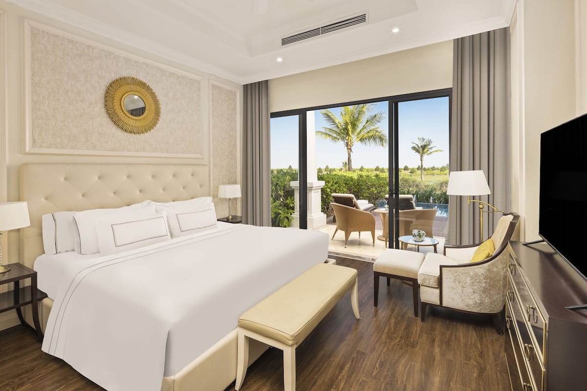 A hotel room with a large bed, a bench, a TV, a dining area with chairs, and a view of palm trees.