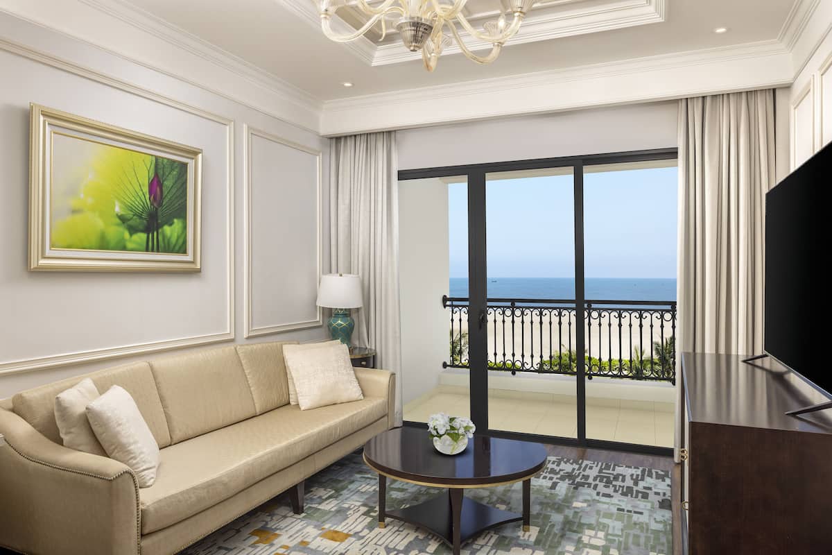 A hotel room with a beige sofa, a small round table with a vase, a painting on the wall, and a large window with a view of the ocean.