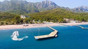 Une zone côtière avec une jetée qui s’avance dans la mer, entourée de montagnes et bordée d’une plage où se trouvent des bâtiments et des arbres.
