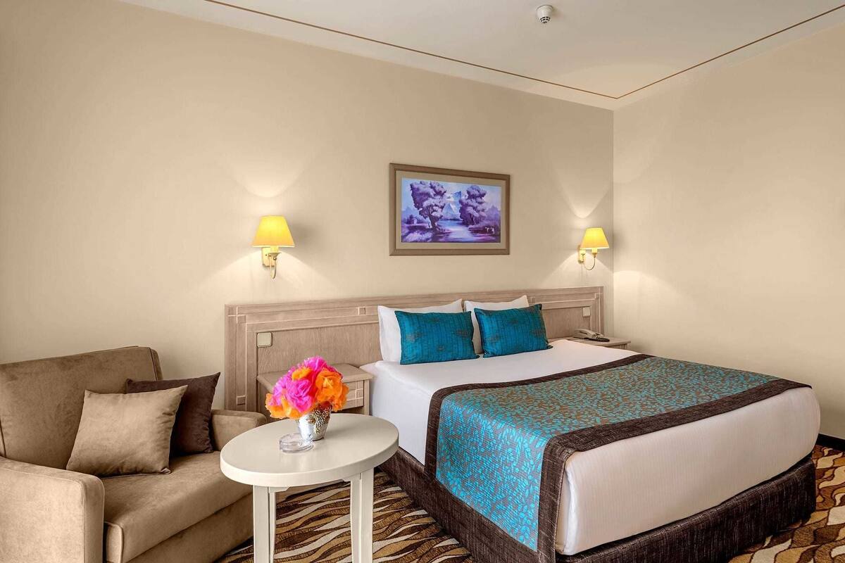A hotel room with a bed, a sofa, a small table with a vase of flowers, and two wall-mounted lamps.