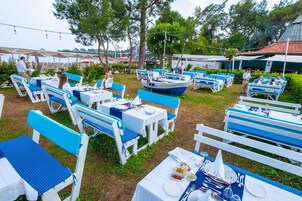 Un área de comedor al aire libre con mesas y sillas dispuestas sobre césped, rodeada de árboles y una masa de agua.