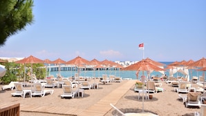 Un complexe hôtelier en bord de mer, avec des rangées de chaises longues à l’ombre de parasols, une promenade en bois et une vue dégagée sur la mer.