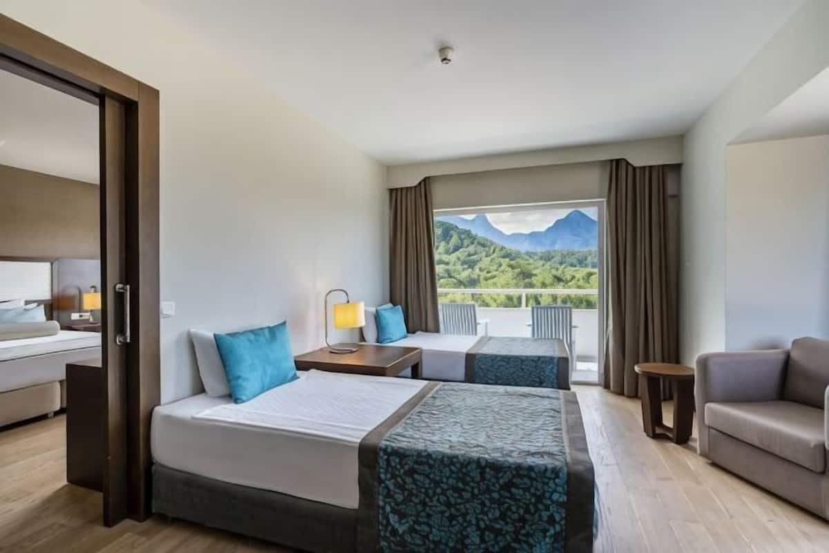 Habitación de hotel con dos camas, un sofá, un balcón con vistas a la montaña y un ventanal grande.