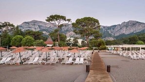 Une plage bordée de rangées de chaises longues et de parasols, une promenade en bois et des montagnes en arrière-plan.