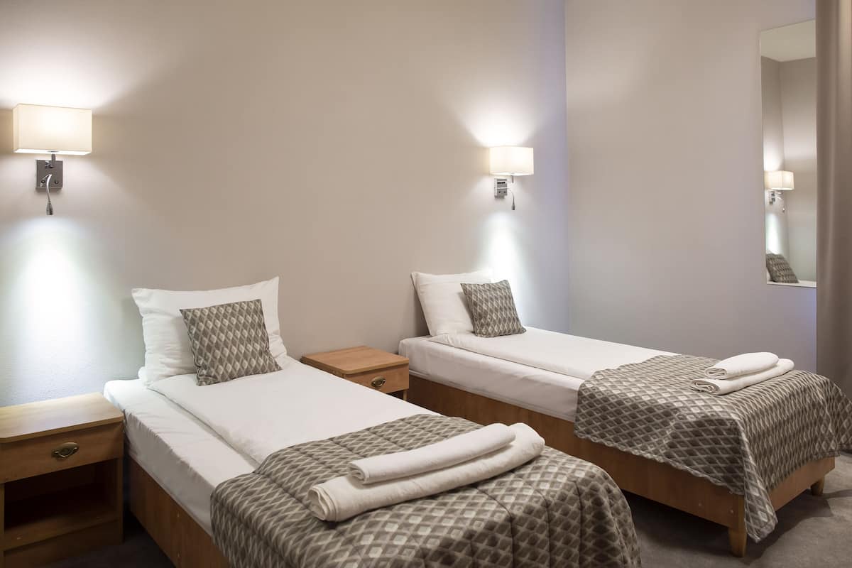 Quarto de hotel com duas camas de solteiro, cada uma com travesseiro e cobertor. Há mesas de cabeceira com abajures e uma gavetinha.