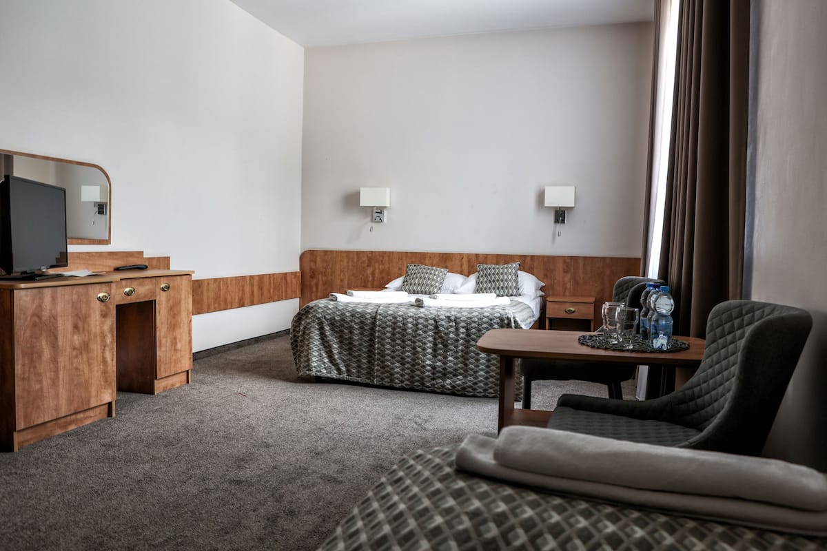Quarto de hotel com cama, escrivaninha com computador, cadeira, mesinha com garrafa de água e televisão.