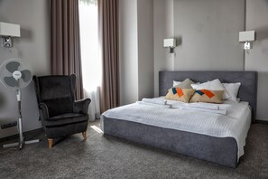 A hotel room with a grey upholstered chair, a bed with white bedding and colorful pillows, a standing fan, and two wall-mounted lamps. - Jan Sander Hotel (Aleksandrow Lodzki)