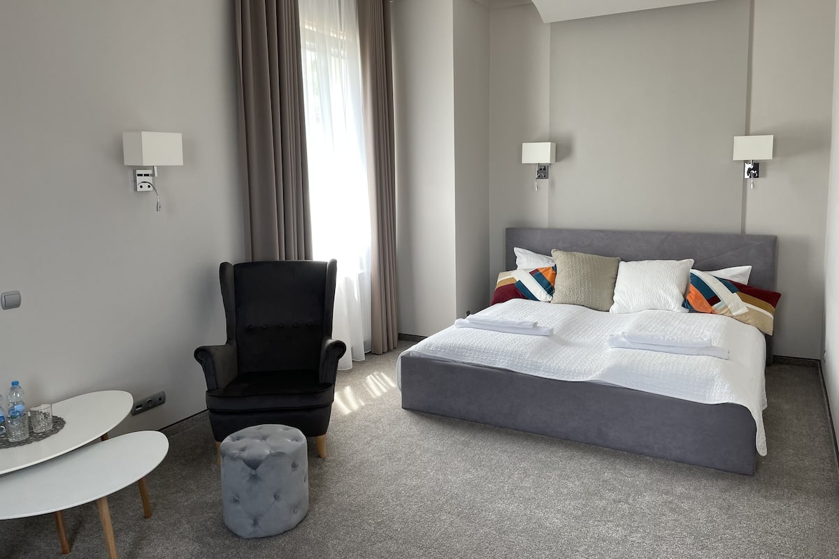 Quarto de hotel com cama, uma cadeira, uma mesa pequena e um banquinho.