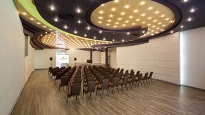 A conference room with rows of chairs and a stage, featuring a modern ceiling design with recessed lighting and a central circular light fixture. - Jan Sander Hotel (Aleksandrow Lodzki)