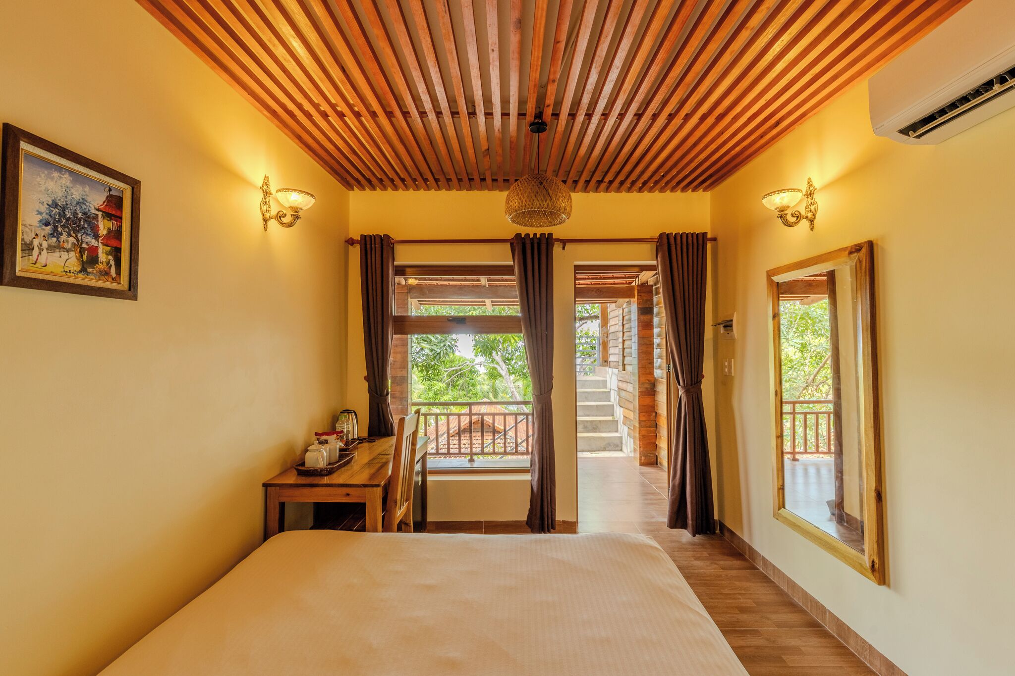 A hotel room with a bed, a desk with a chair, a window with curtains, a painting on the wall, and a staircase leading to an outdoor area.
