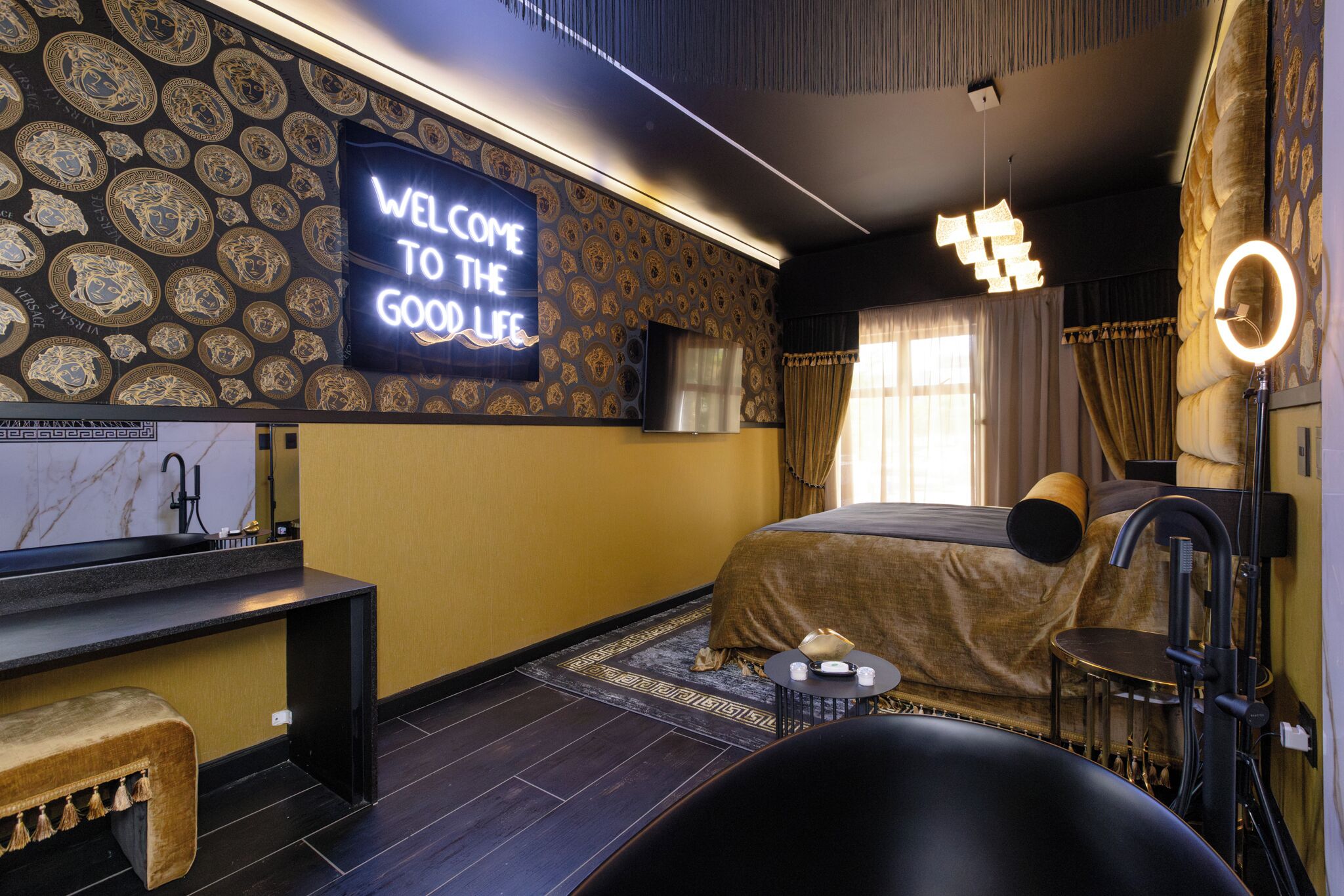 Une chambre à coucher moderne, avec un lit, une coiffeuse, un banc et un téléviseur affichant le message : « BIENVENUE DANS UN MONDE DE PLAISIRS ».