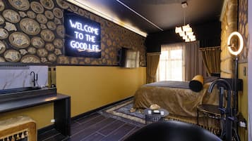 Un dormitorio moderno con cama, tocador, banqueta y televisión mostrando “WELCOME TO THE GOOD LIFE”.