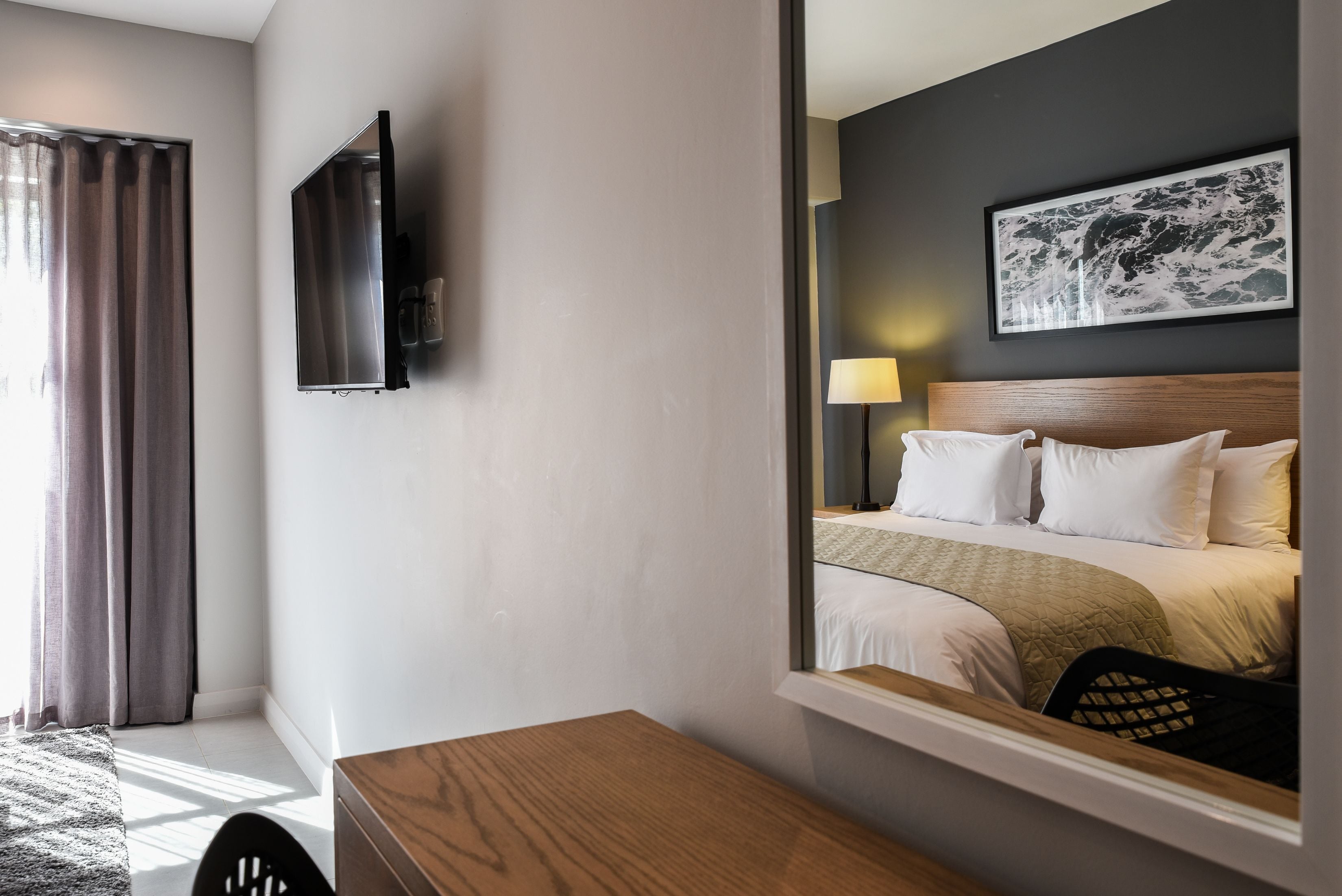 A hotel room with a bed, a TV mounted on the wall, a window with curtains, and a desk with a chair.