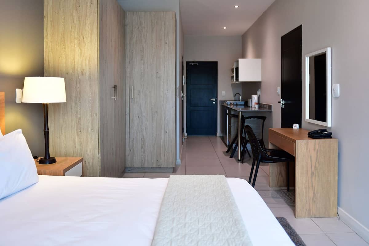 A hotel room with a bed, bedside lamp, desk, chair, and a small kitchenette.