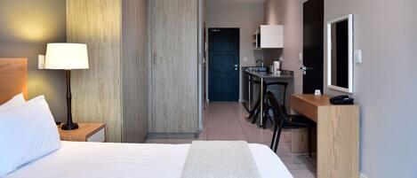 Habitación de hotel con cama, lámpara de noche, escritorio, silla y una kitchenette pequeña.