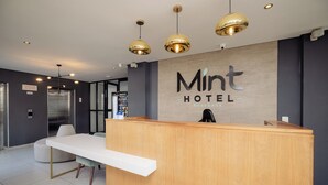 A hotel reception area with a wooden reception desk, a white counter, and a sign that reads "Mint Hotel Rosebank".