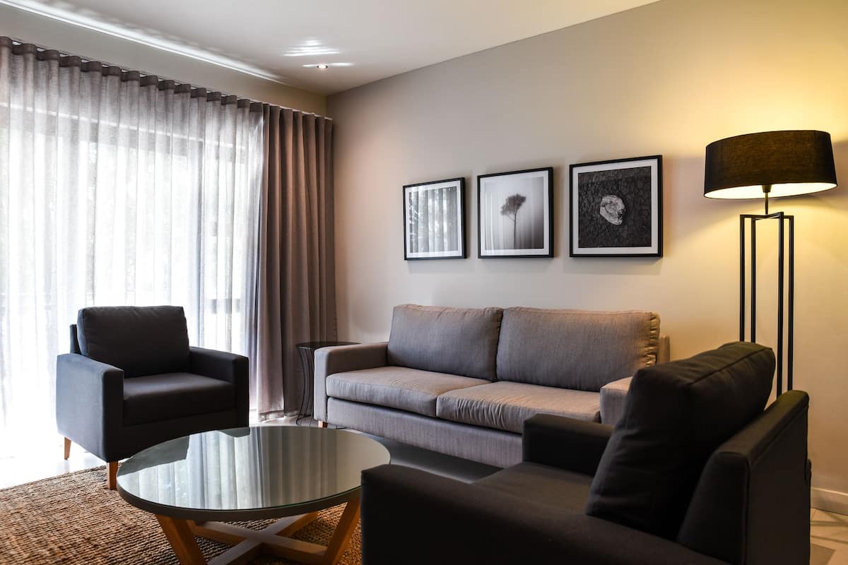 A modern living room with a grey sofa, a black armchair, and a glass coffee table. There are three framed artworks on the wall and a floor lamp.