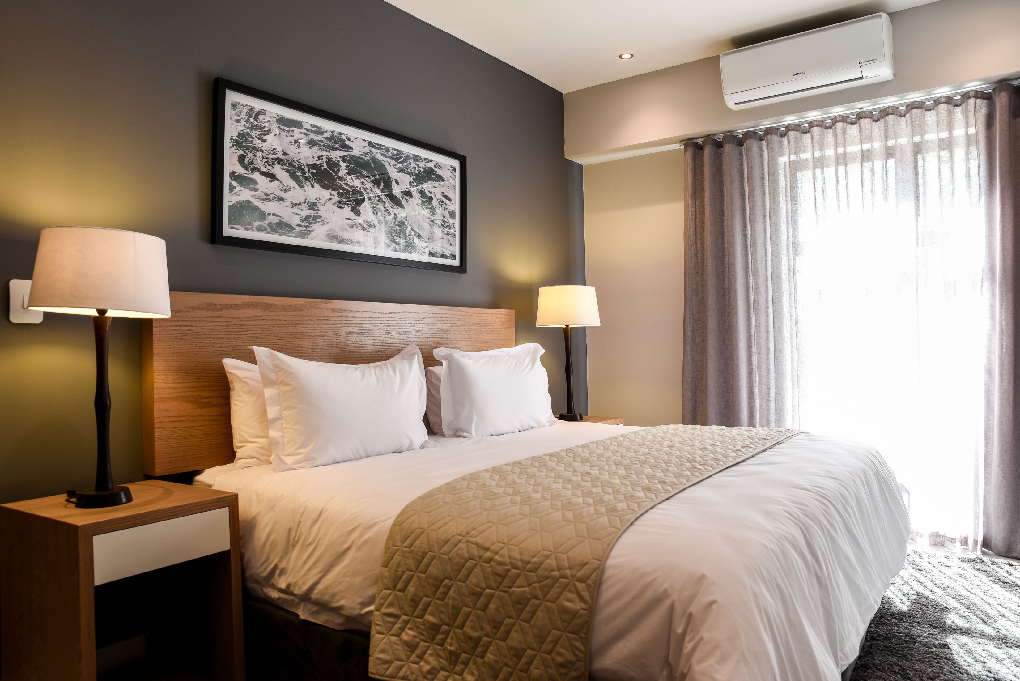 A hotel room with a large bed, two bedside lamps, a framed artwork on the wall, and an air conditioning unit.
