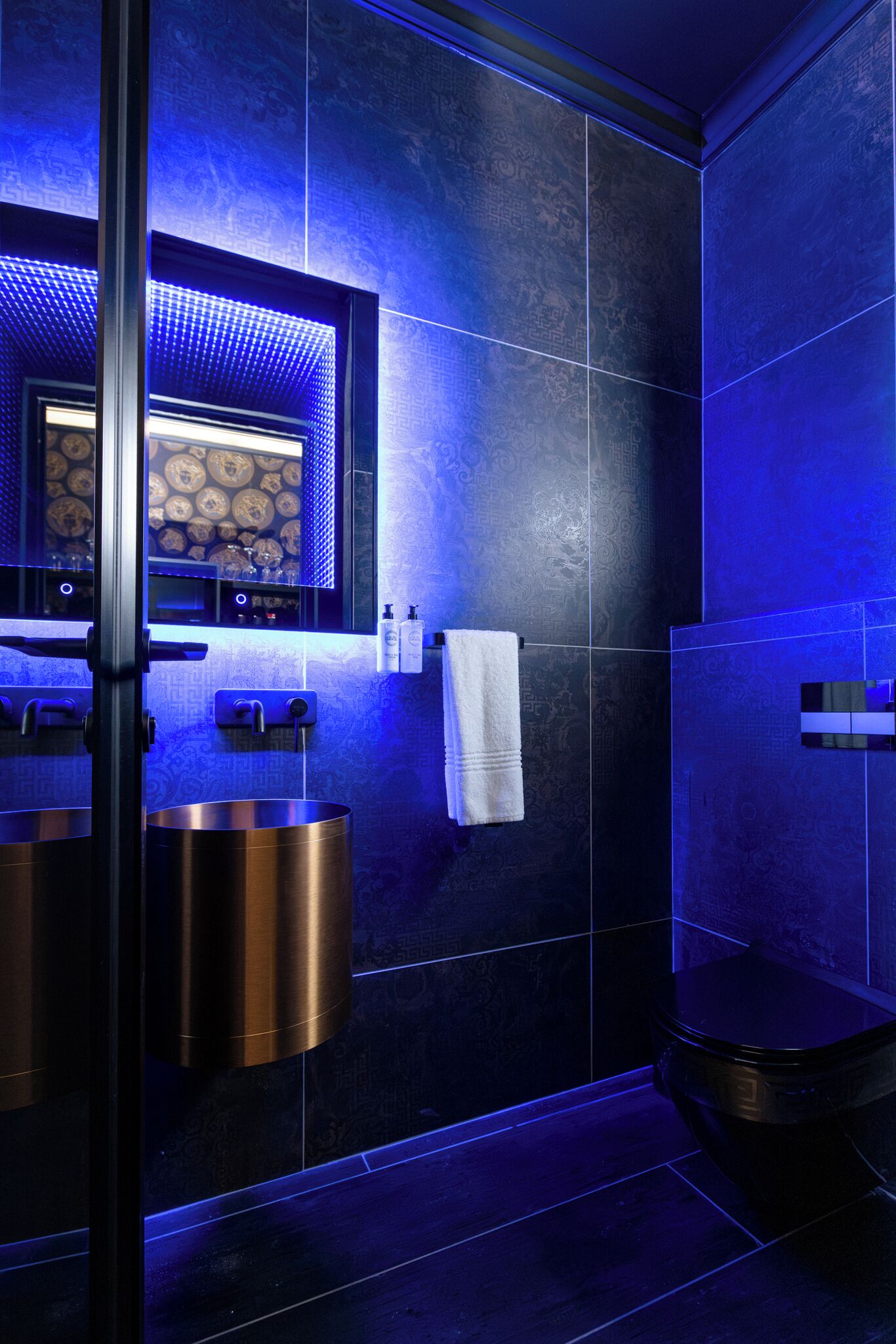 A modern bathroom with dark blue tiles, a large mirror, a round sink, and a toilet.